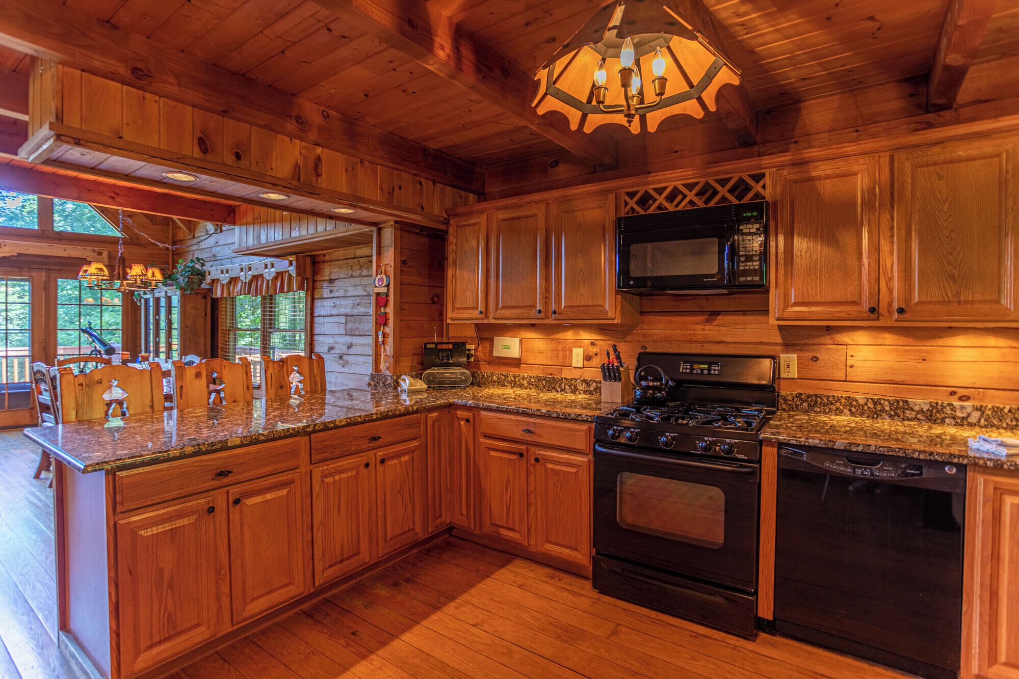 A wooden cabin-style kitchen with a granite countertop, wooden cabinets, a stove, microwave, and a chandelier.
