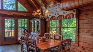 A wooden dining room with a table and chairs, a chandelier, and large windows with blinds. - Apple of my Eye (Banner Elk)