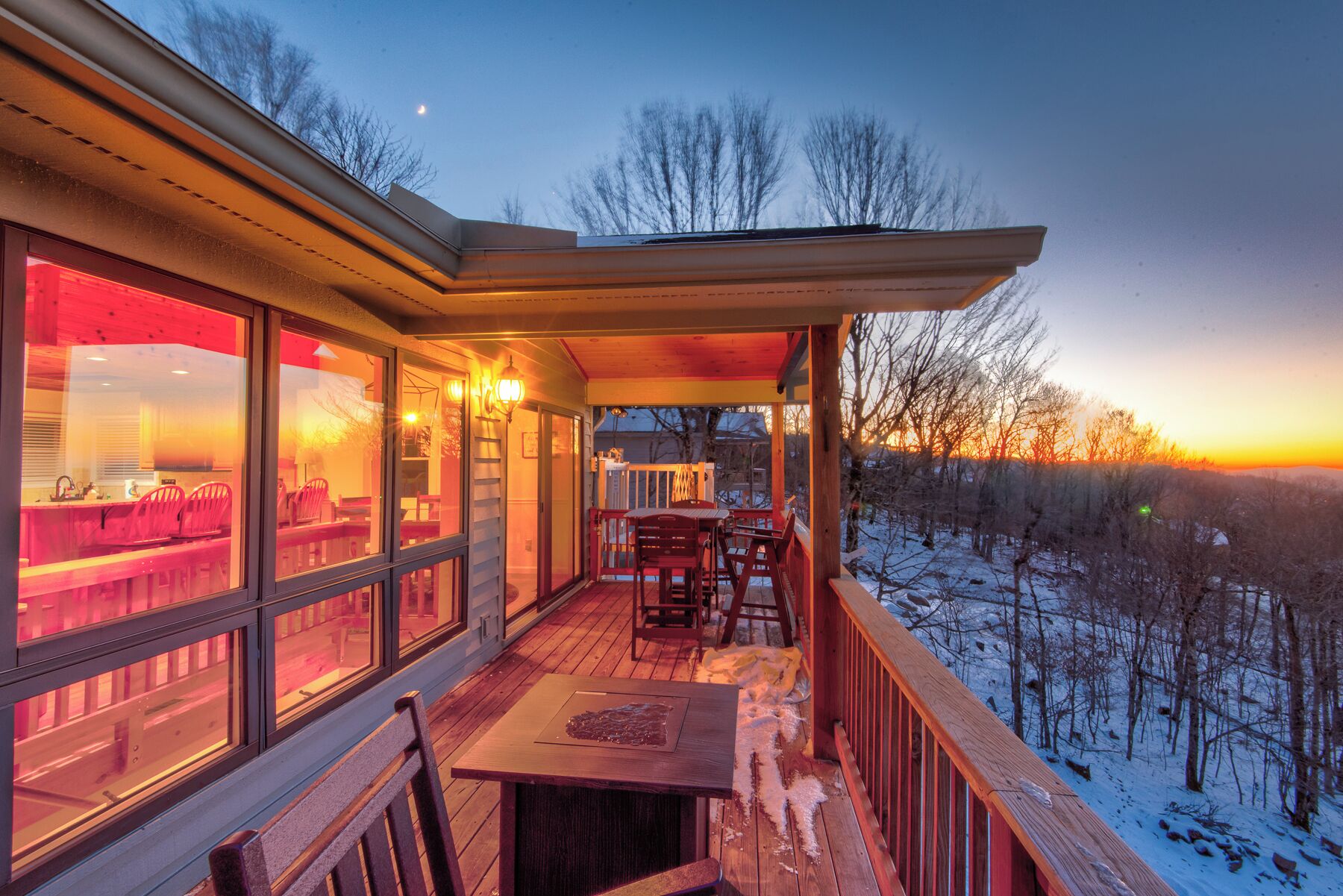 A wooden deck with a table and chairs, overlooking a snowy landscape at sunset.