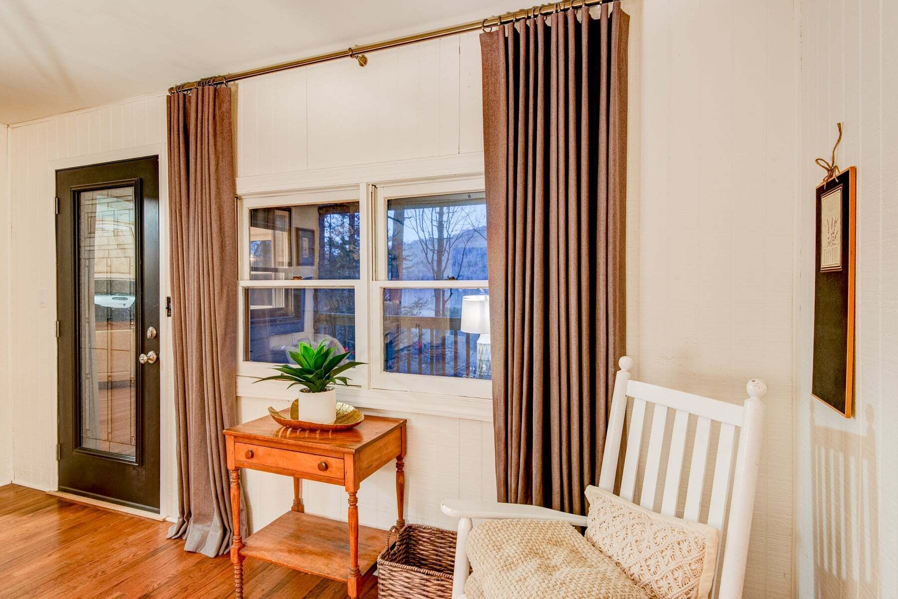 A room with a wooden floor, a white chair, a small table with a plant, a window with curtains, and a door.