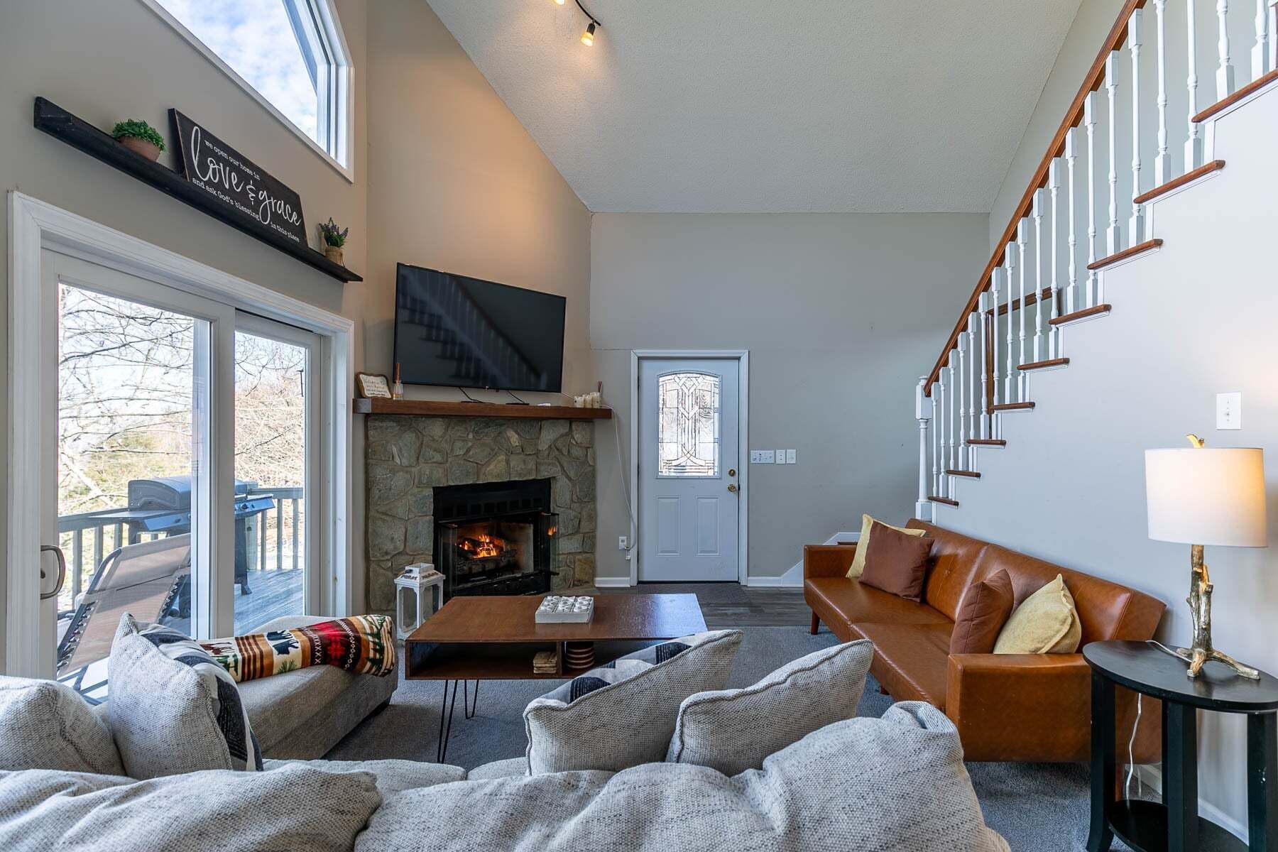 Una sala de estar con chimenea de piedra, un televisor montado encima, un sofá, una mesa de centro y una escalera con barandillas blancas.