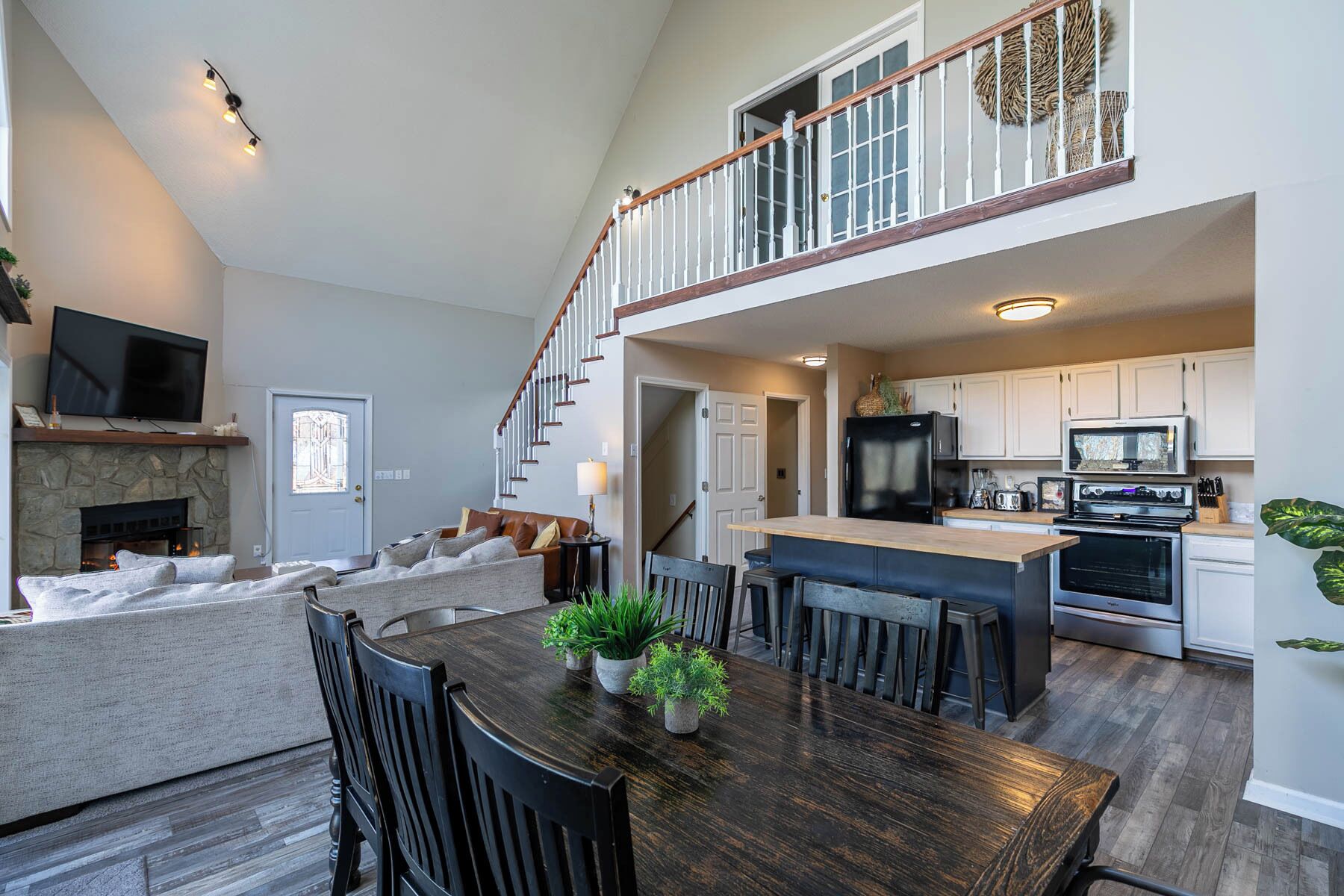 A spacious living area with a fireplace, a dining table with chairs, a kitchen with modern appliances, and a staircase leading to an upper level.