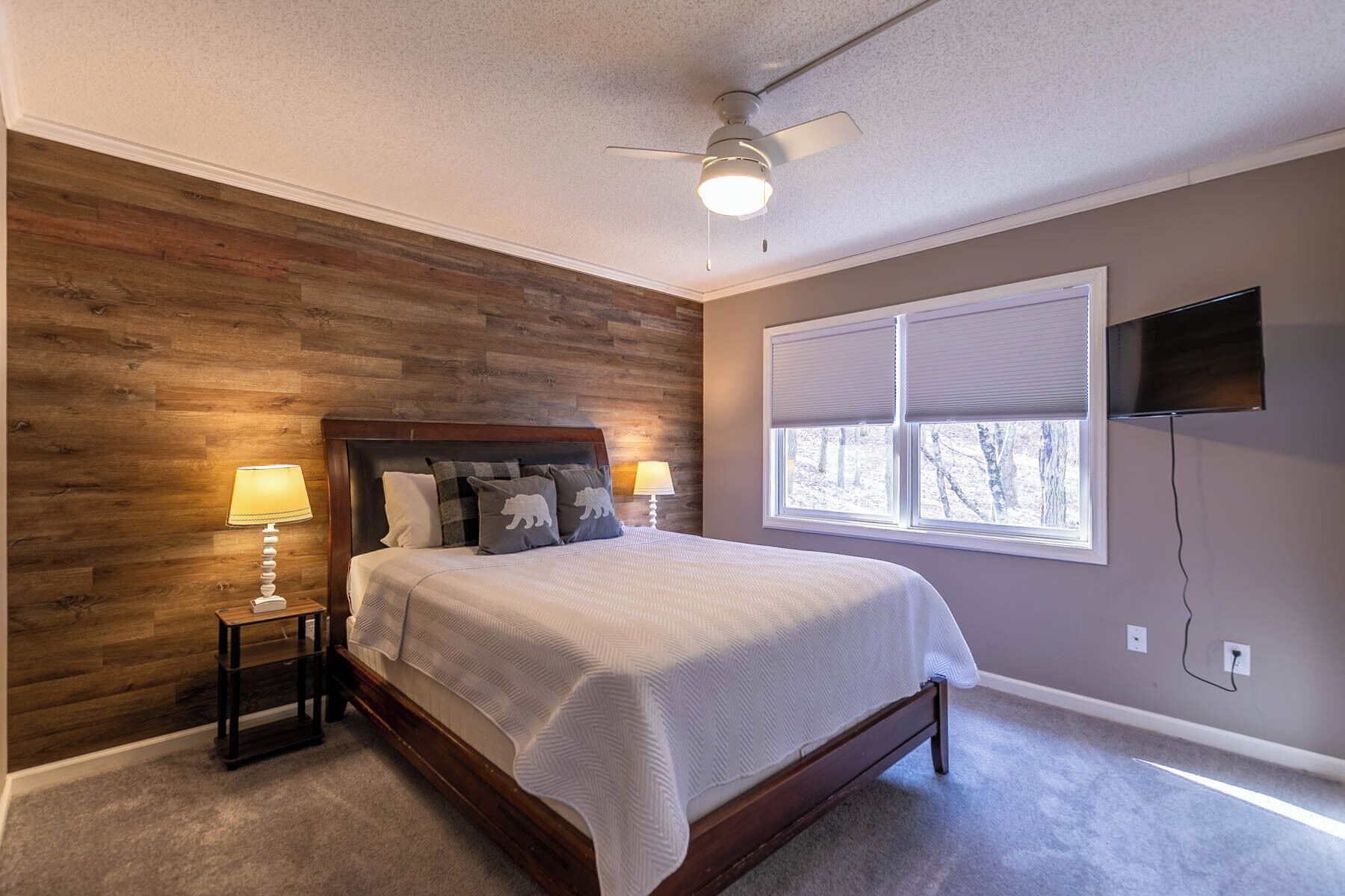 Une chambre avec un mur en bois, un lit recouvert d’une courtepointe blanche, une table de chevet avec des lampes, un ventilateur de plafond et une fenêtre avec des stores.