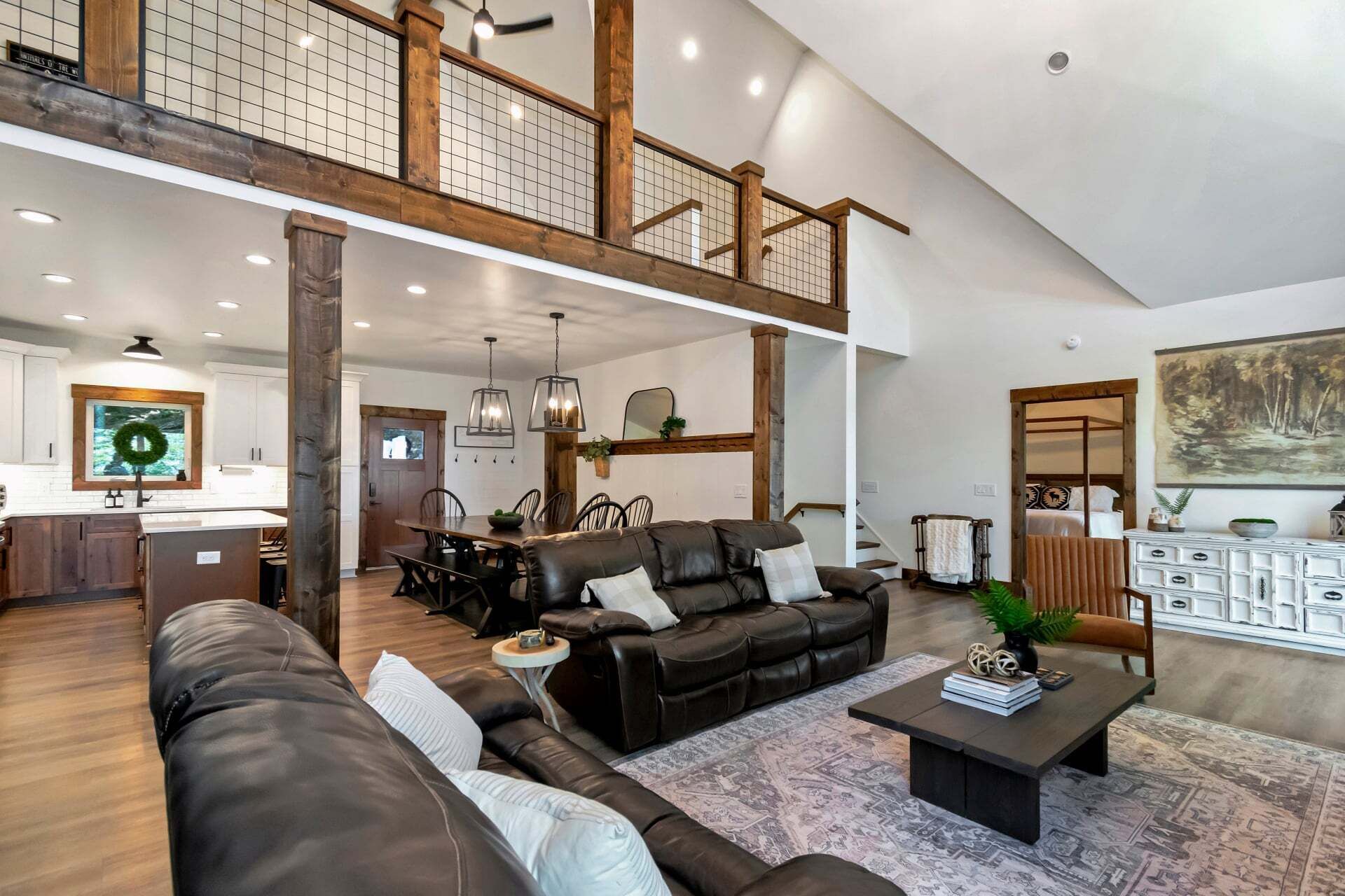 A spacious living area with a high ceiling, a dining area with a wooden table and chairs, and a bedroom with a large bed and a painting.