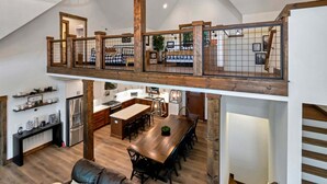 A spacious loft with a high ceiling, exposed wooden beams, a mezzanine level with a railing, a kitchen with modern appliances, and a dining area with a large wooden table. - Beech Blessings - Luxury Spacious Beech Mtn Home With Huge Views (Banner Elk)