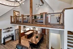 Ein geräumiger Loft mit hoher Decke, freiliegenden Holzbalken, einer erhöhten Zwischenebene mit Geländer, einer Küche mit modernen Geräten und einem Essbereich mit großem Holztisch.
