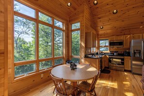 Eine Holzhütte mit einem runden Esstisch und Stühlen, großen Fenstern, einer Küche mit modernen Geräten und Blick auf den Wald.