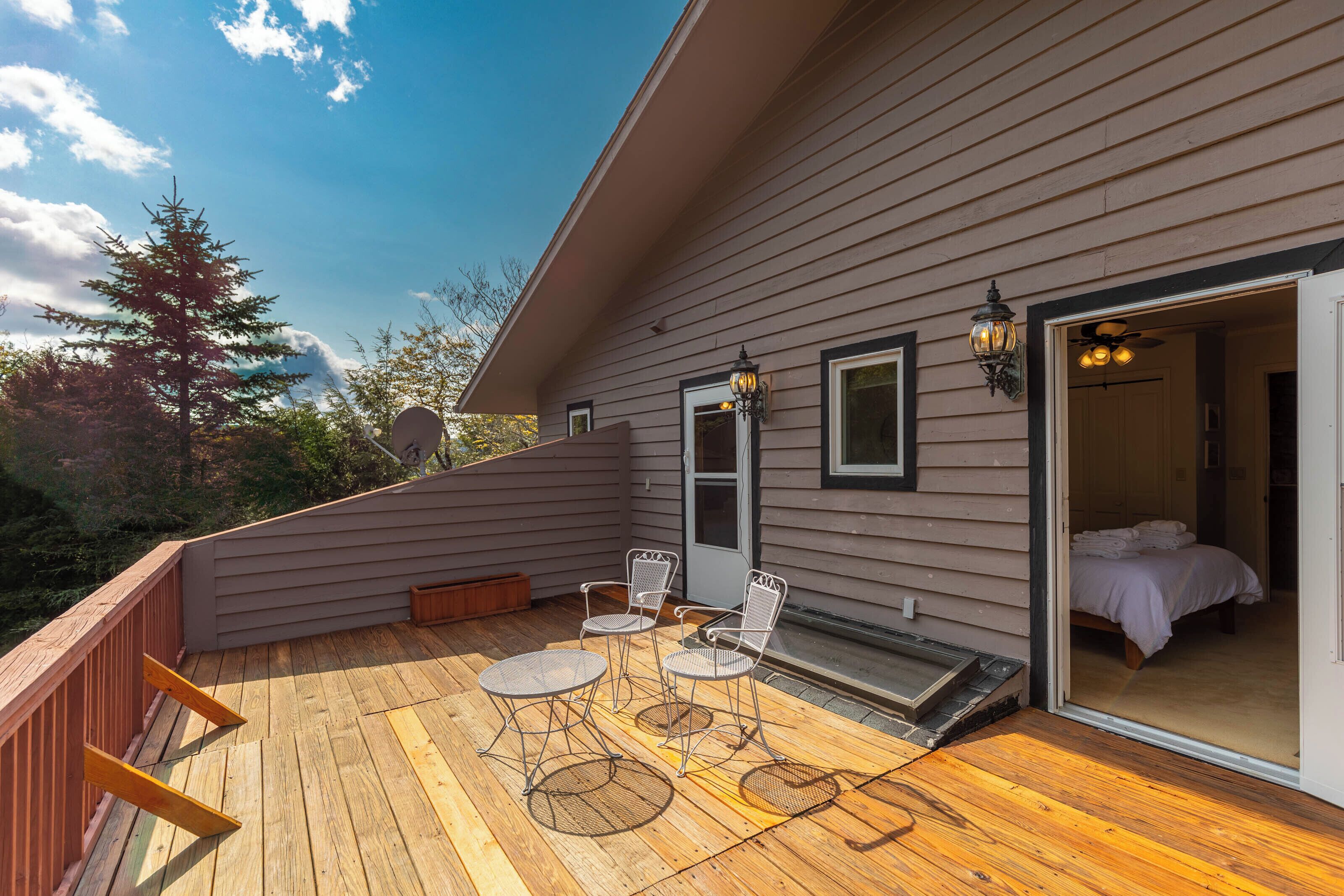 Una terraza de madera con mobiliario exterior, una puerta corrediza de vidrio hacia un dormitorio y una antena parabólica.