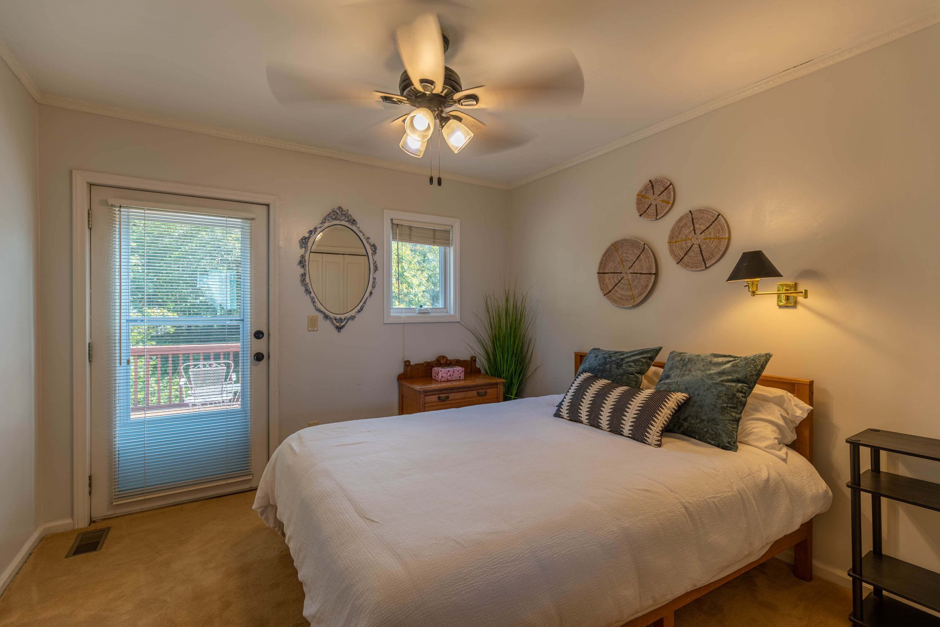 A bedroom with a bed, a ceiling fan, a window with blinds, a door, a small table with a plant, and wall decor.