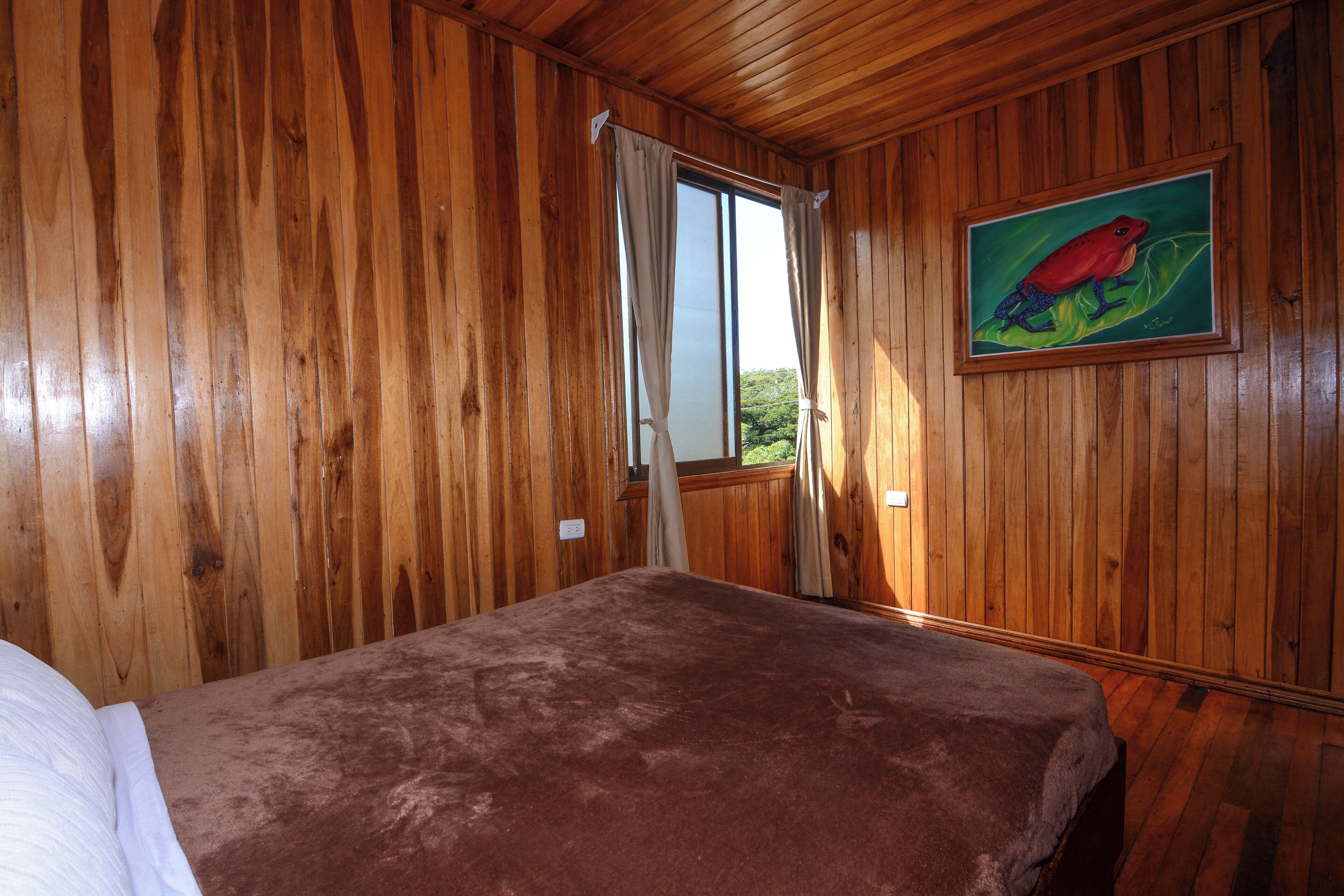 A wooden-paneled room with a bed, a window with curtains, and a painting of a frog on the wall.