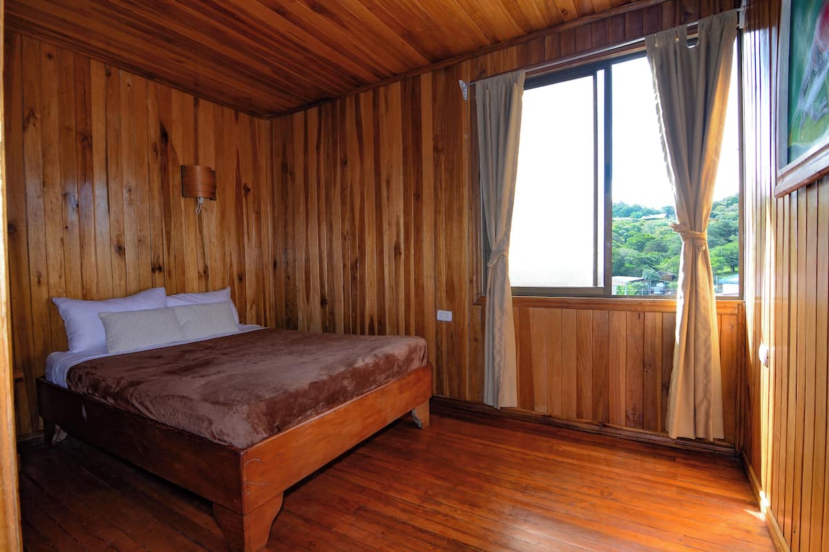 Una habitación con paneles de madera, una cama, un ventanal con cortinas y vistas a la vegetación.