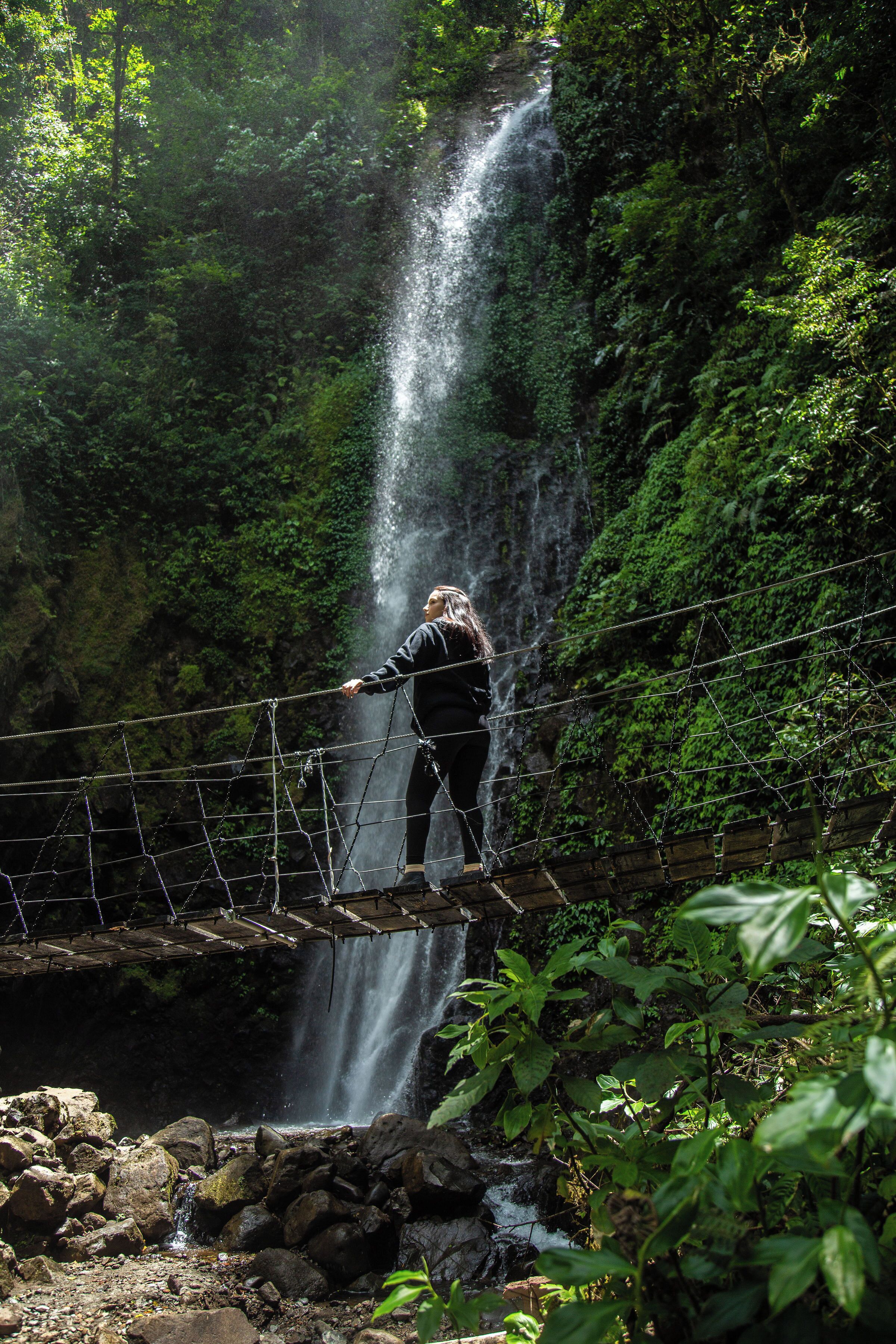 Una persona de pie sobre un puente colgante sobre una cascada en un área boscosa.