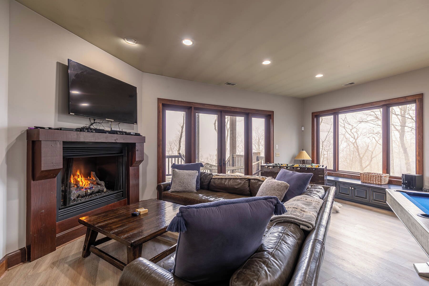A spacious living room with a fireplace, leather sofa, wooden coffee table, and a large window overlooking a deck.