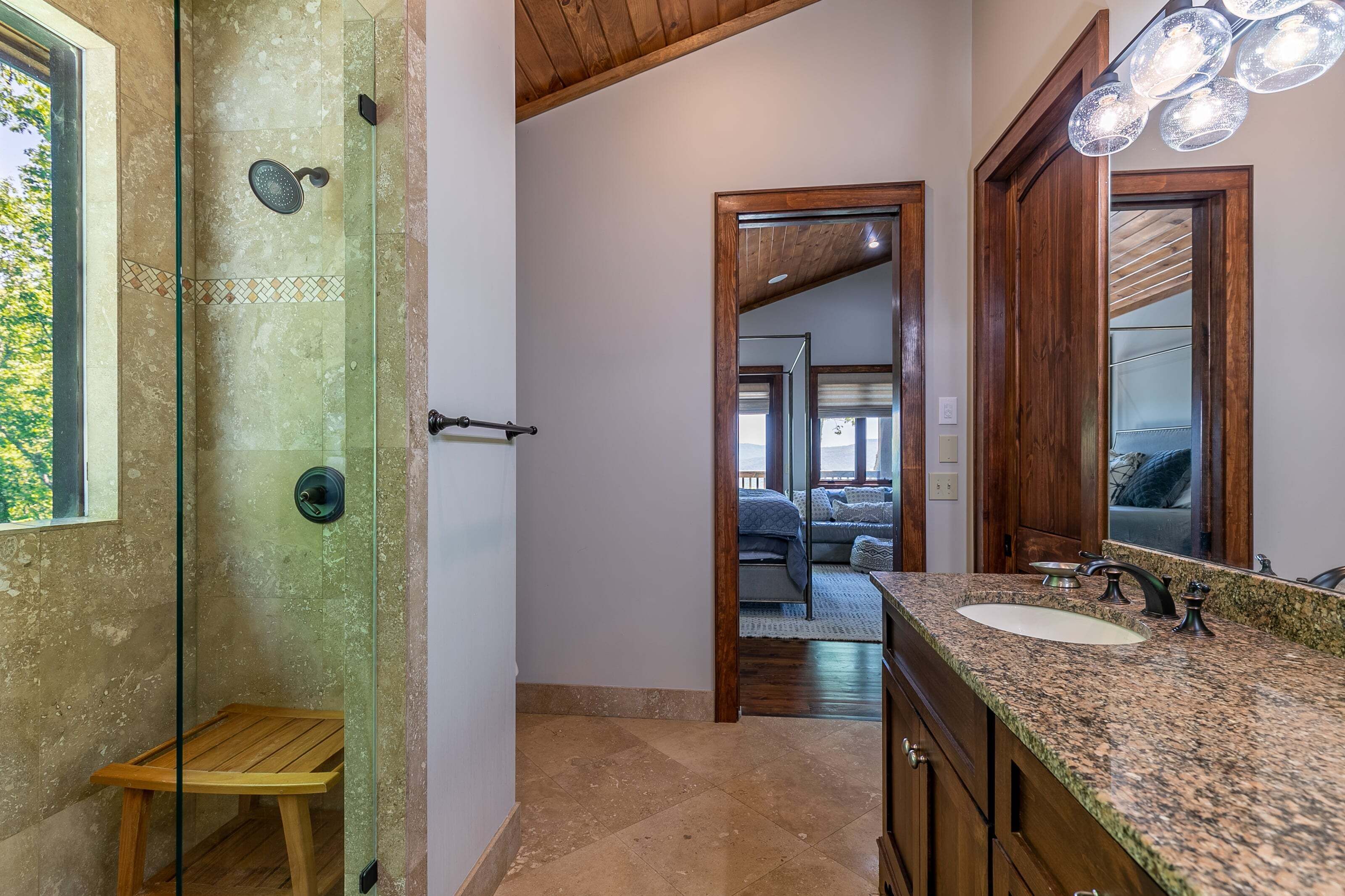 A bathroom with a glass shower enclosure, wooden bench, granite countertop, and a large mirror.
