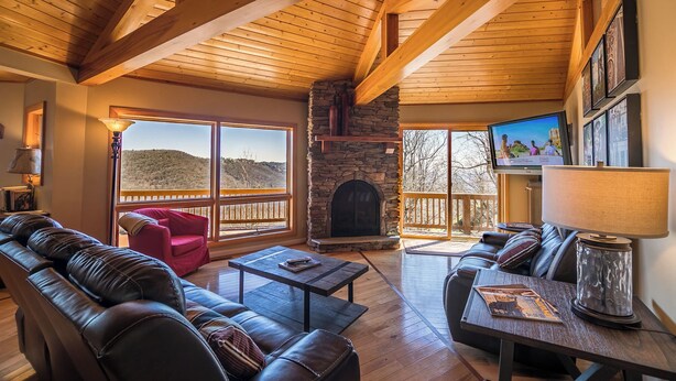 Ein geräumiges Wohnzimmer mit Kamin aus Stein, Ledersofas, einem Fernseher und großen Fenstern mit Blick ins Freie.