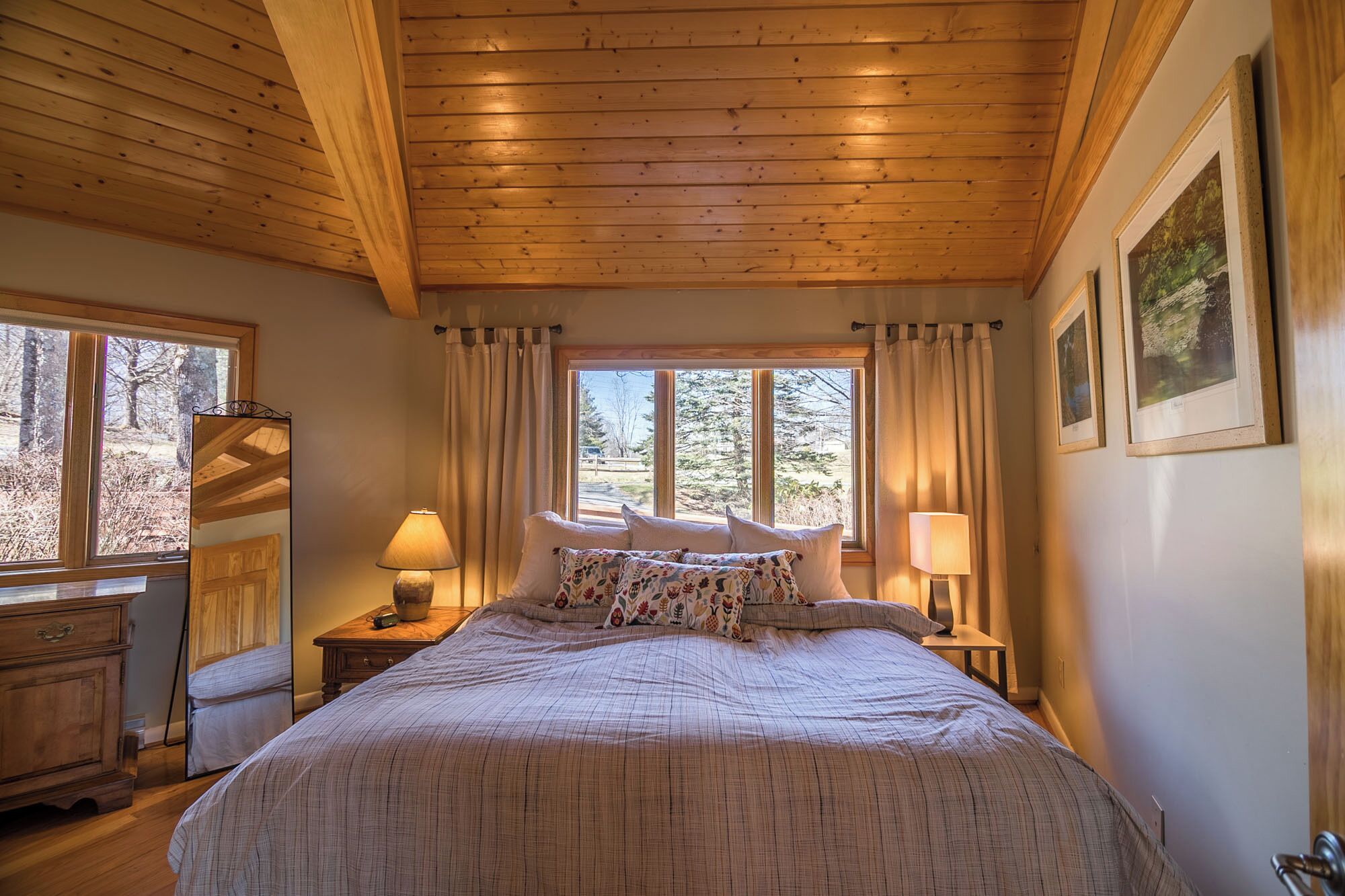 A bedroom with a wooden ceiling, a large bed with patterned pillows, two bedside tables with lamps, a dresser, and a window with curtains.