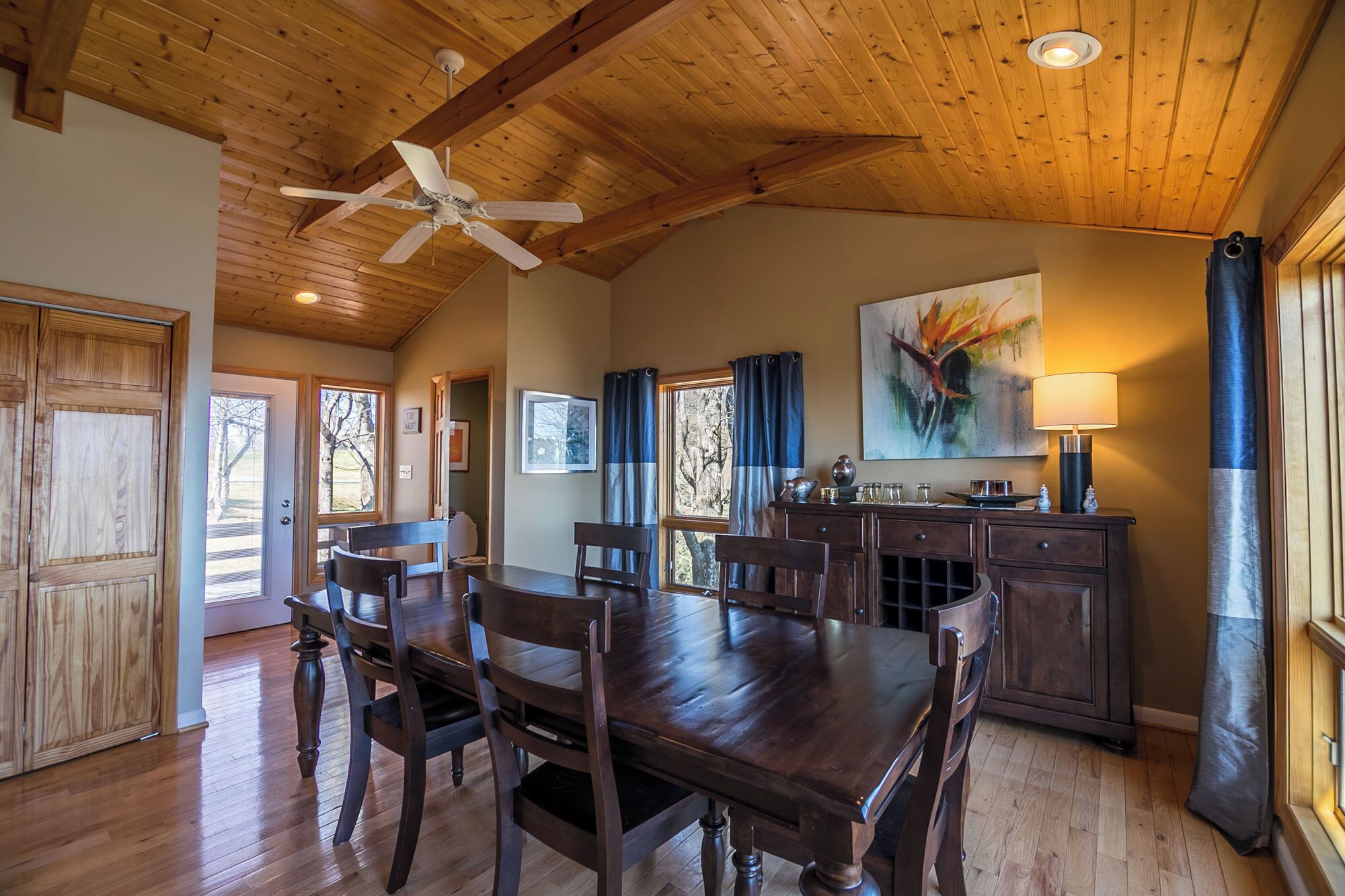 A dining area with a wooden table and chairs, a ceiling fan, and a painting on the wall.