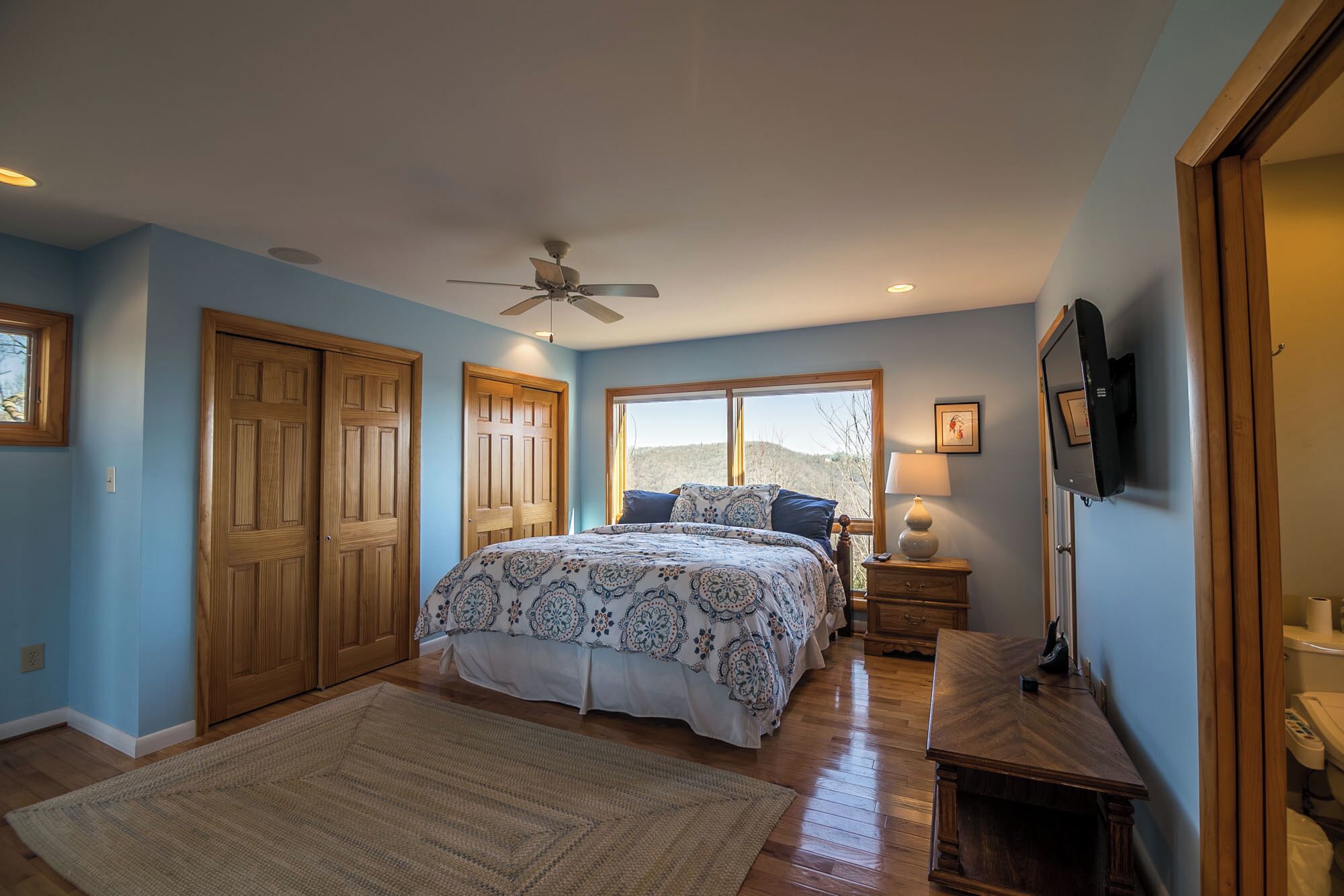 A bedroom with a large bed, wooden wardrobe, ceiling fan, TV, and a view of the outdoors.