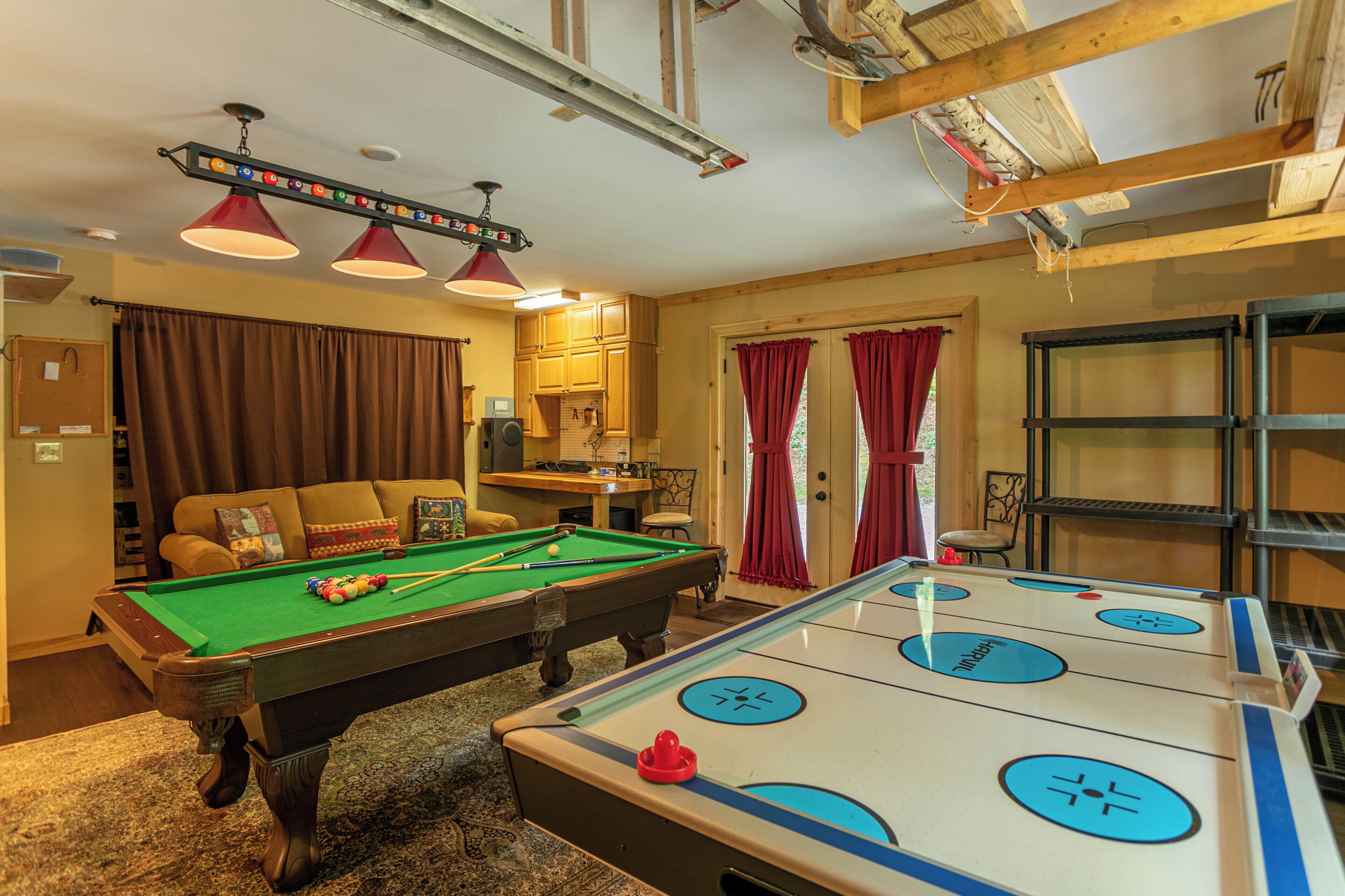 A pool table, a billiards table, a sofa, and a bookshelf in a room with wooden beams and a ceiling fan.