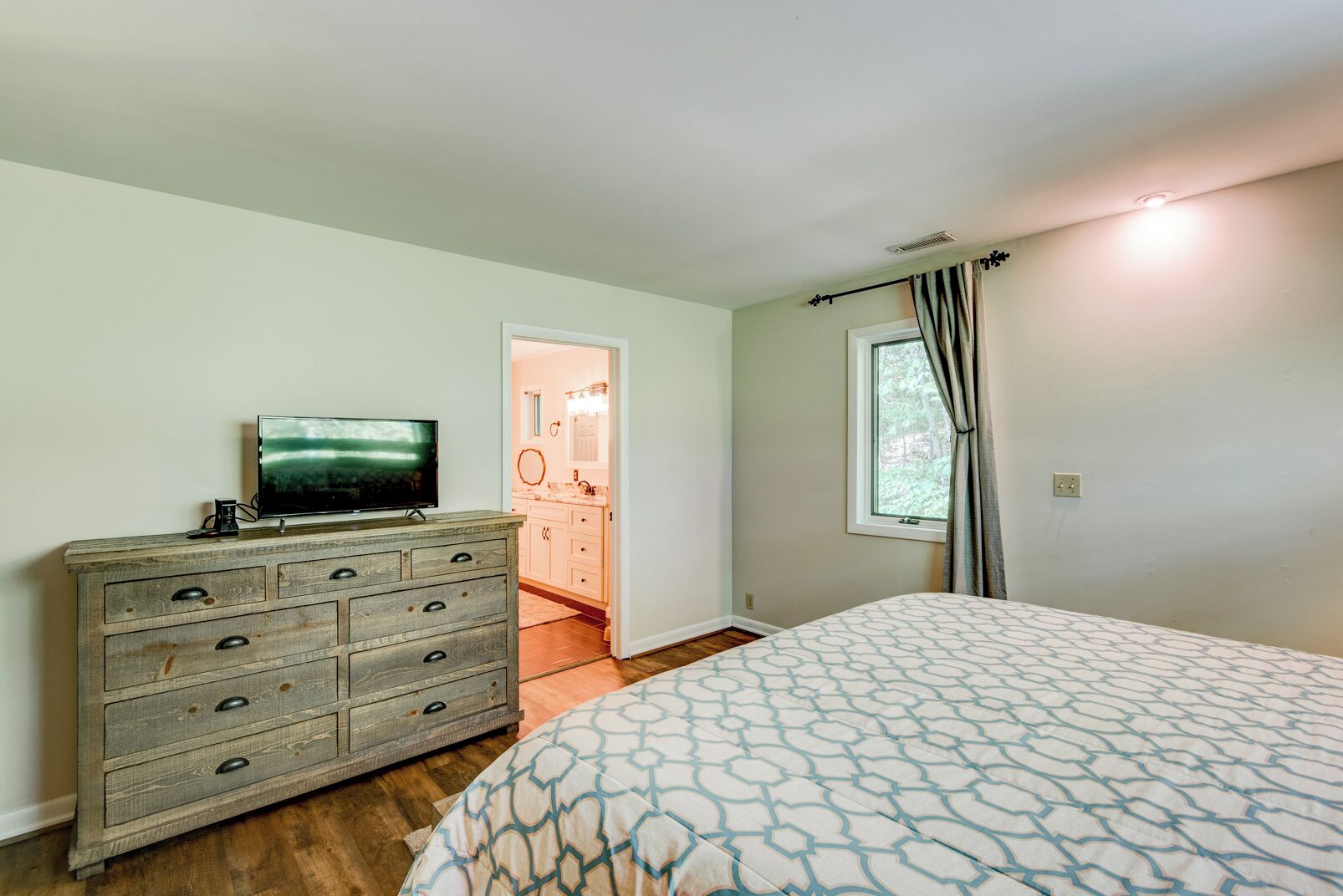 Une chambre avec une commode en bois, un lit avec une literie à motifs, un téléviseur et une salle de bain visible à travers une porte ouverte.