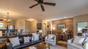 A spacious living room with a dark brown leather sofa, a light grey armchair, a wooden coffee table, and a ceiling fan.