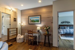 Una habitación con pared de madera, un escritorio con lámpara, una silla, una mesita de noche con un florero, y una cama con colcha azul y blanca a cuadros.