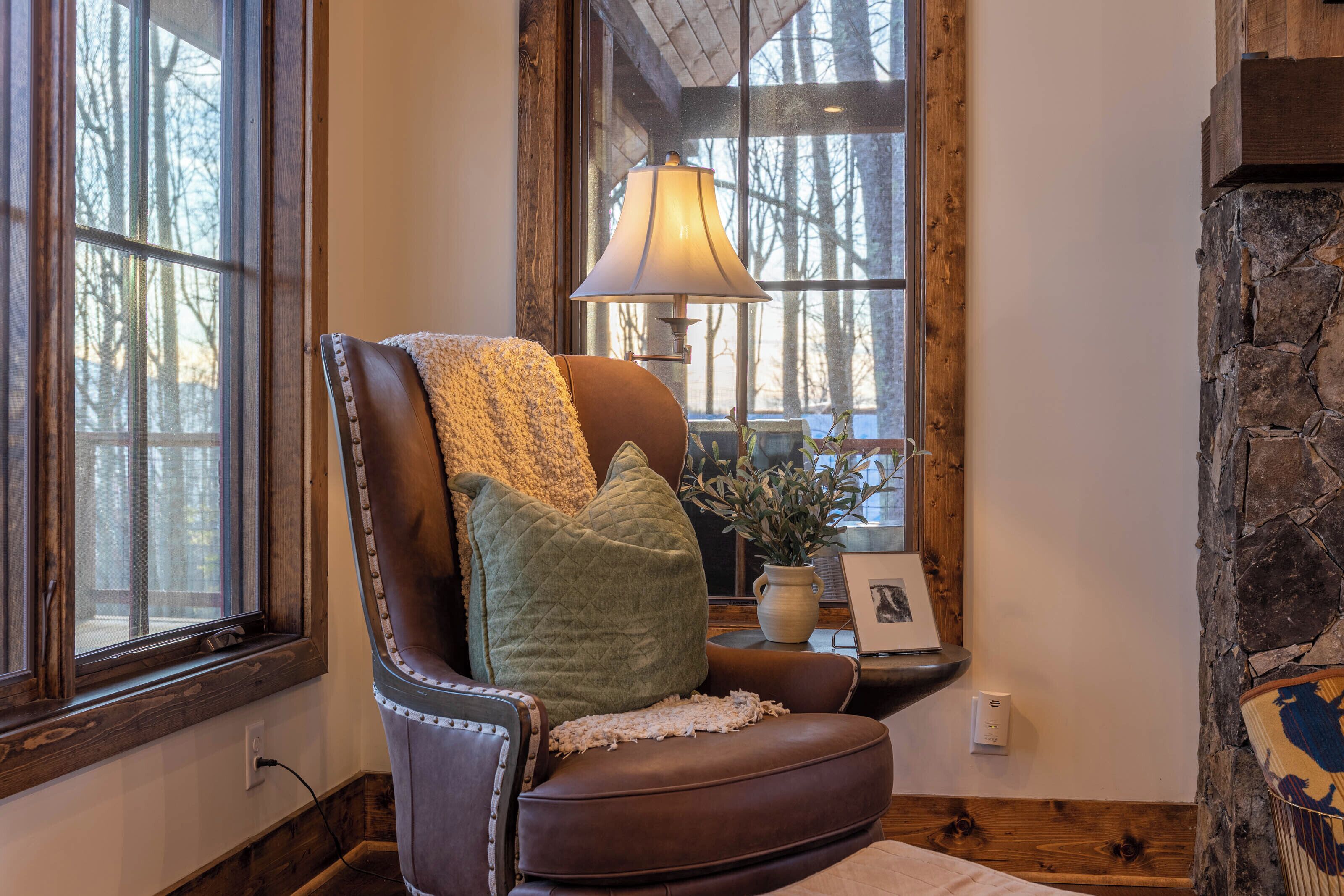 A leather armchair with a quilted pillow and a lamp, positioned near a window with a view of trees.