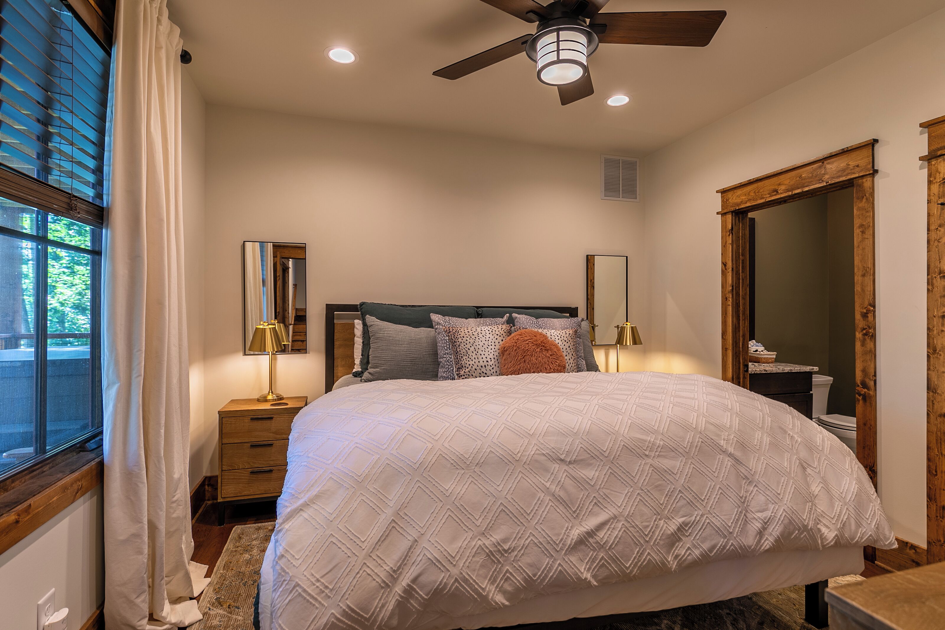 A bedroom with a bed, a ceiling fan, a window with curtains, a nightstand, a mirror, and a wooden door.