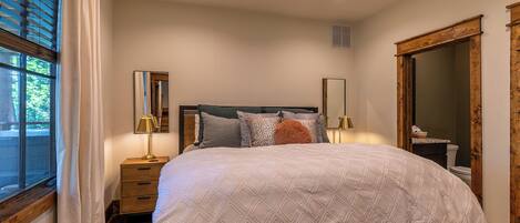 A bedroom with a bed, a ceiling fan, a window with curtains, a nightstand, a mirror, and a wooden door.