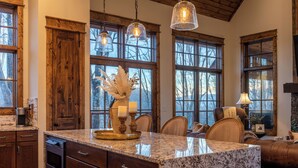A kitchen with granite countertops, wooden cabinets, and a large island. There are pendant lights, a fireplace, and a view of trees through large windows. - Tipsy Elk at Eagles Nest (Banner Elk)