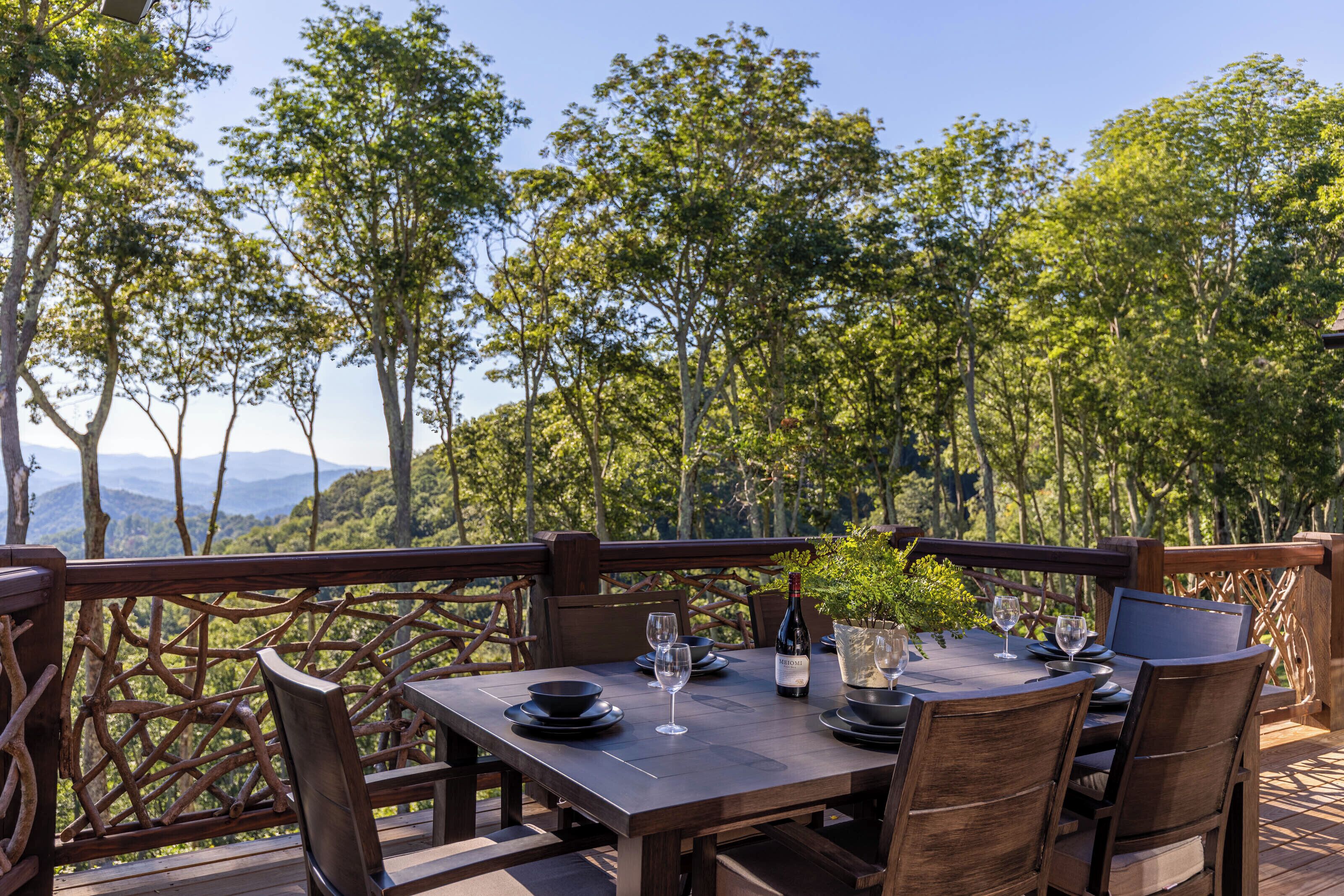 Un espace de restauration en plein air avec une table dressée pour deux personnes, entouré de chaises en bois, offrant une vue sur un paysage boisé avec des montagnes à l’horizon.