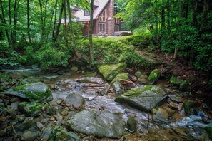 Ein WaldBach mit Steinen, ein Holzhäuschen im Hintergrund und eine Steinbrücke.