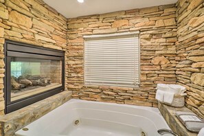 A bathroom with a stone wall, a bathtub, a window with blinds, and a fireplace. - Lillys Mountain Escape (Banner Elk)