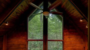 L’intérieur d’un chalet en bois, avec une grande fenêtre, un ventilateur de plafond et une vue sur une forêt.