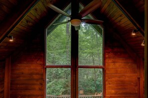 Innenansicht einer Holzhütte mit großem Fenster, Deckenventilator und Blick auf einen Wald.