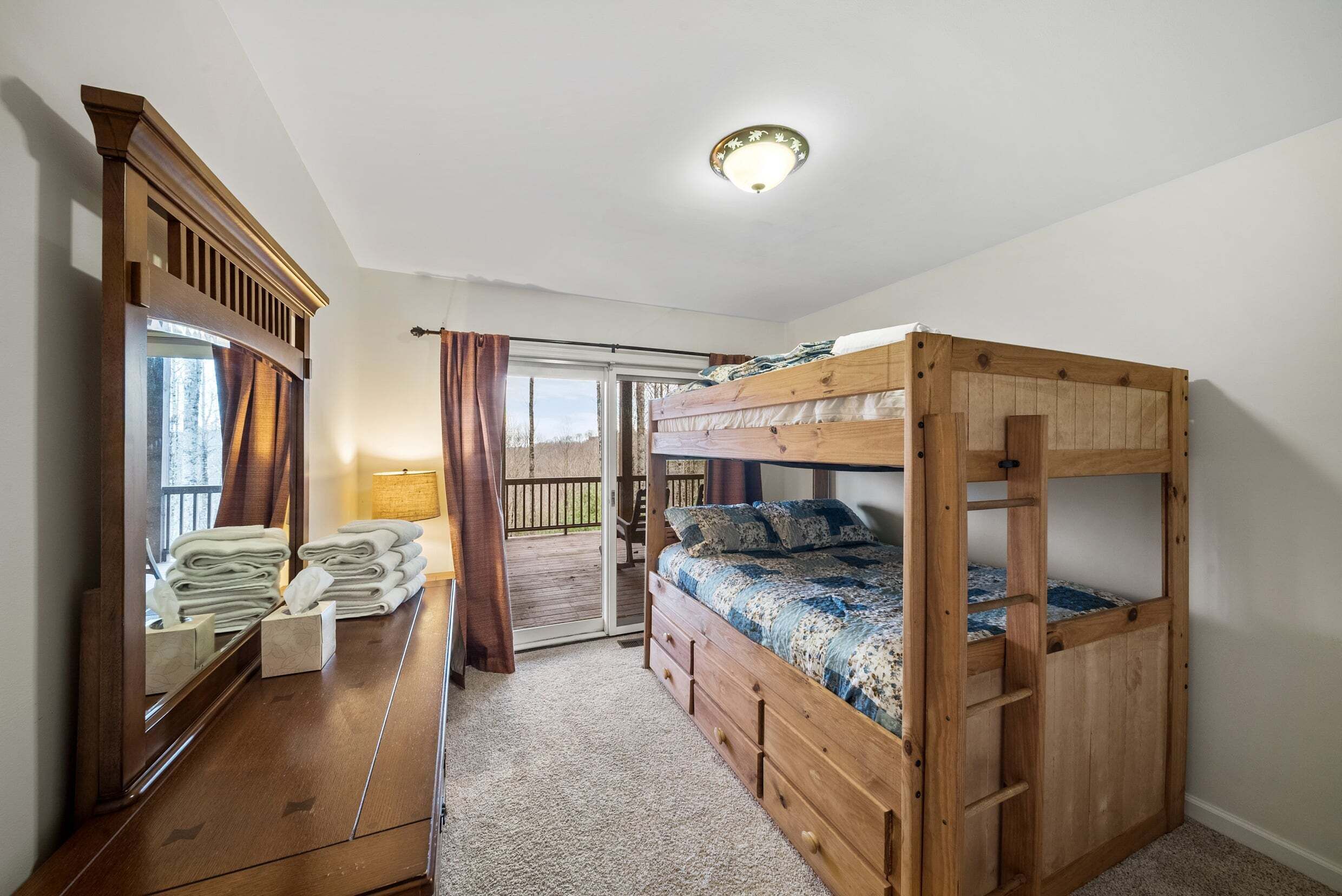 A bunk bed room with a wooden headboard, a mirror, a wooden dresser, and a view through a sliding door to an outdoor deck.