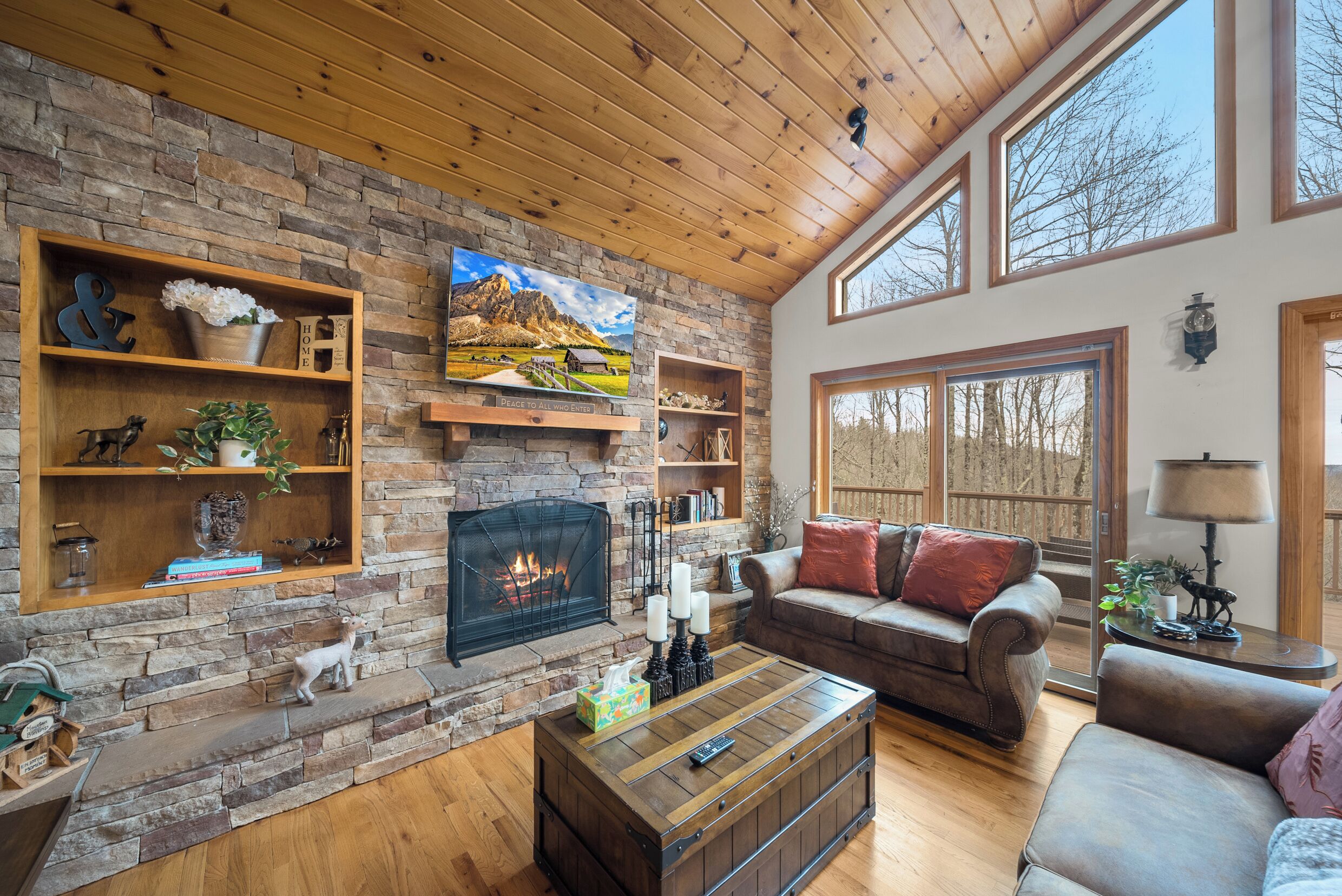 A cozy living room with a stone fireplace, wooden shelves, a sofa, and a large window.