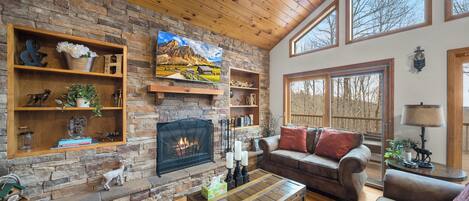 A cozy living room with a stone fireplace, wooden shelves, a sofa, and a large window.