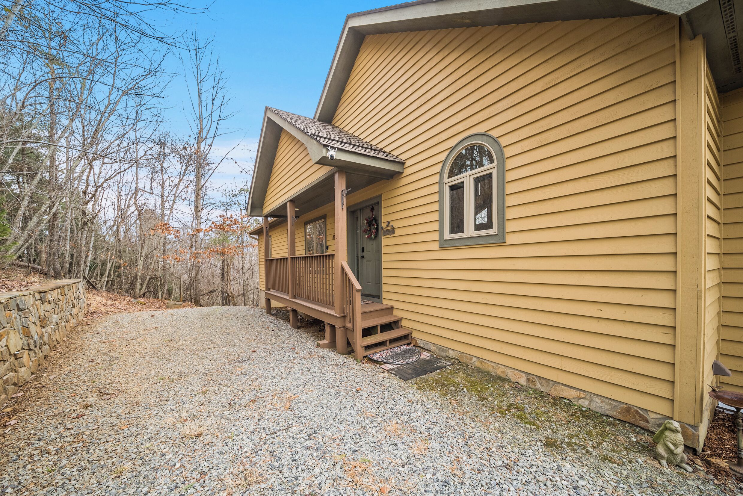 A yellow wooden house with a porch, a stone wall, and a gravel driveway.