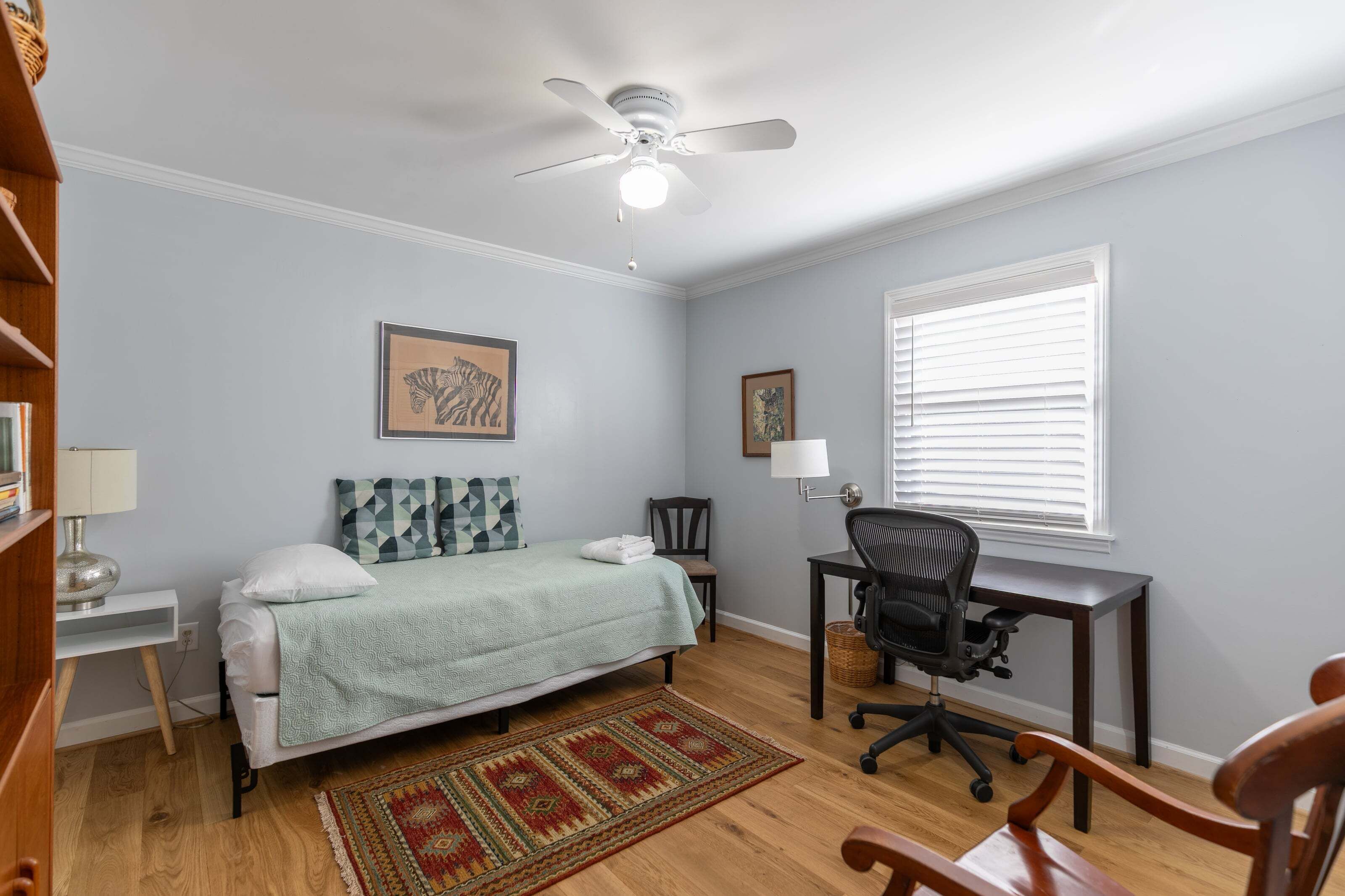 A bedroom with a bed, desk, chair, ceiling fan, and a window with blinds.