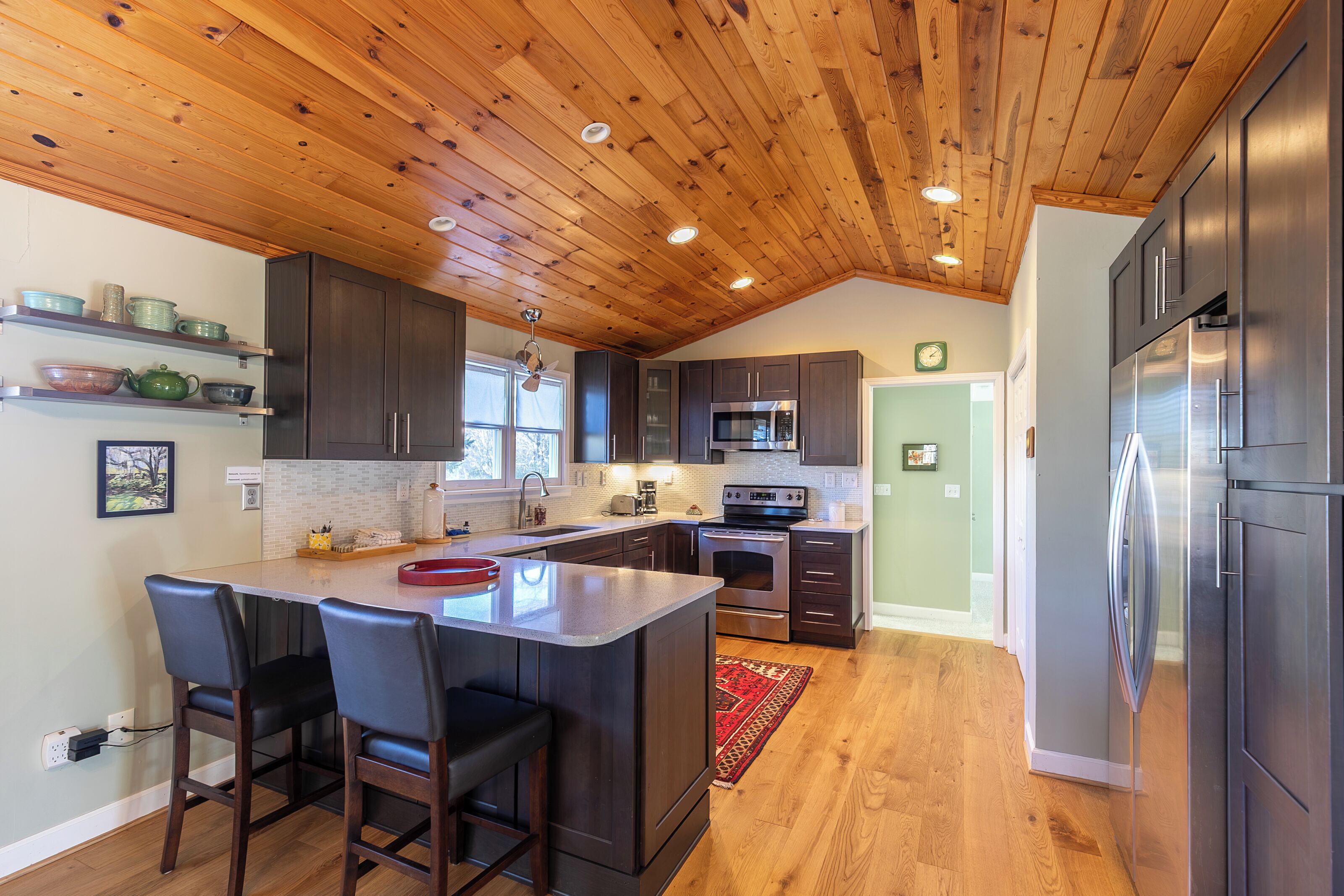 A kitchen with wooden ceiling, dark cabinets, stainless steel appliances, and a central island with bar stools.