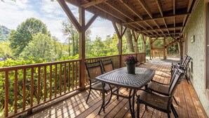A covered outdoor deck with wooden railings, a table with chairs, and a view of a forested area. - Moose Preserve - Cabin Near Valle Crucis With hot Tub, Pool Table, & Firepit (Sugar Grove)