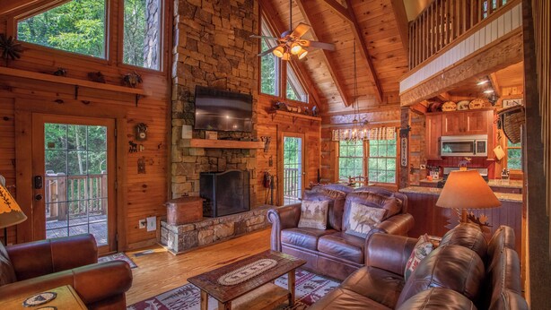 A log cabin interior with a stone fireplace, leather sofas, a television, and a kitchen area. - Moose Preserve - Cabin Near Valle Crucis With hot Tub, Pool Table, & Firepit (Sugar Grove)