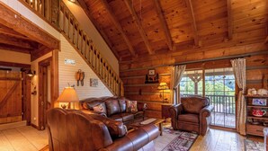A log cabin interior with a wooden staircase, leather furniture, and a large window offering a view of the outdoors. - Moose Preserve - Cabin Near Valle Crucis With hot Tub, Pool Table, & Firepit (Sugar Grove)