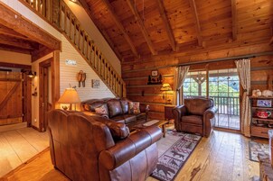 Interior de una cabaña de troncos con escalera de madera, mobiliario de cuero y un ventanal con vistas al exterior.