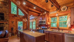 A rustic cabin-style kitchen with a large island, wooden cabinets, and a stone fireplace. - Moose Preserve - Cabin Near Valle Crucis With hot Tub, Pool Table, & Firepit (Sugar Grove)