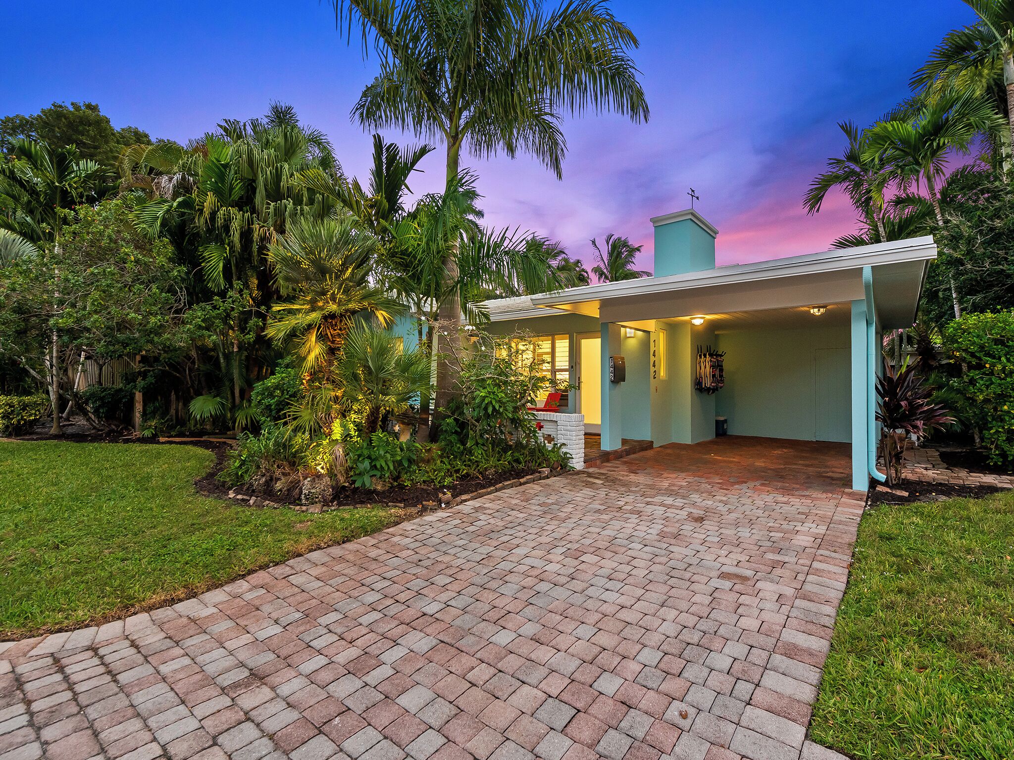 A house with a paved driveway, surrounded by lush greenery and palm trees.