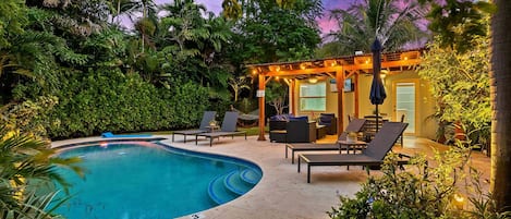 Una piscina con sillones de descanso, una pérgola con luces colgantes y un jardín bien cuidado.