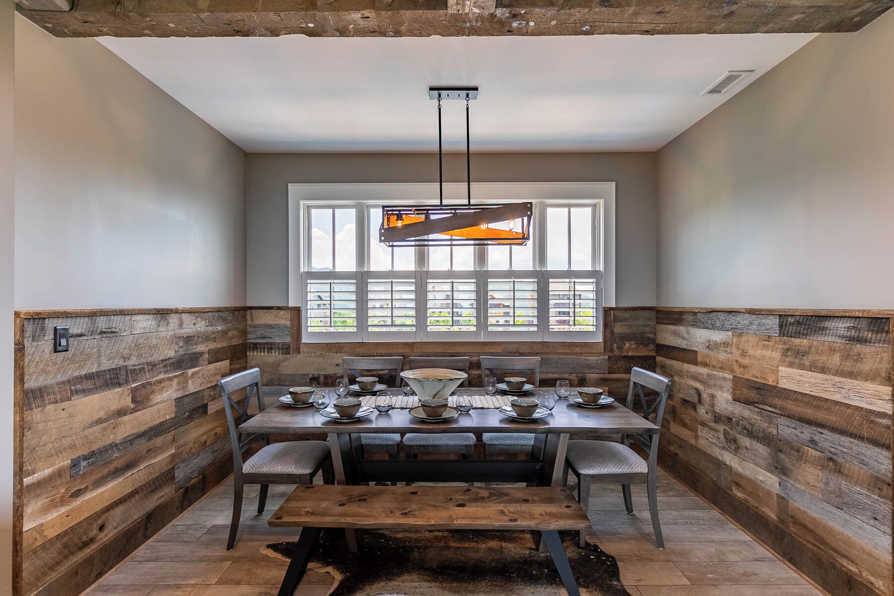 Un comedor con una mesa de madera preparada para cuatro personas, un banco y un gran ventanal con persianas.