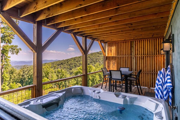 A wooden deck with a hot tub, outdoor seating, and a view of a forested mountain range. - Mountain Kandy at Blue Ridge Mountain Club (Lenoir)